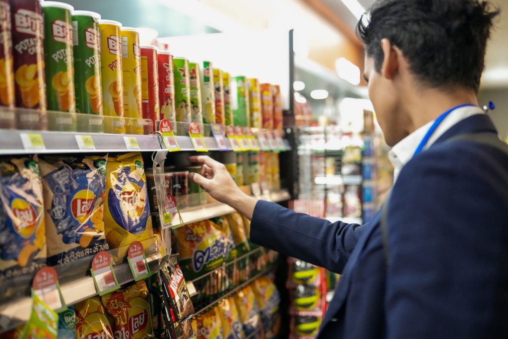 An Asian office worker in formal wear picking up a snack after work in a well stocked shop aisle. Sunlit, uncluttered framing that supports text and brand elements.